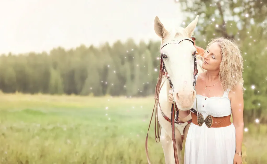 Mandy Magro in a white dress with a brown belt cuddles a white horse in a grassy field with trees in the background.